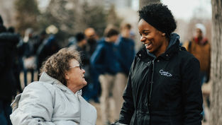 Two women converse outdoors, one in a wheelchair with a gray coat, the other smiling in a black "Christ in the City" jacket; blurred crowd behind.
