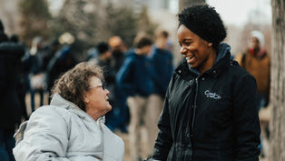 Two women converse outdoors, one in a wheelchair with a gray coat, the other smiling in a black "Christ in the City" jacket; blurred crowd behind.