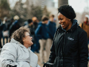 Two women converse outdoors, one in a wheelchair with a gray coat, the other smiling in a black "Christ in the City" jacket; blurred crowd behind.