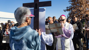 A priest in white robes and sunglasses prays before a large cross outdoors. A crowd, some with prayer books, stands solemnly under a clear sky.
