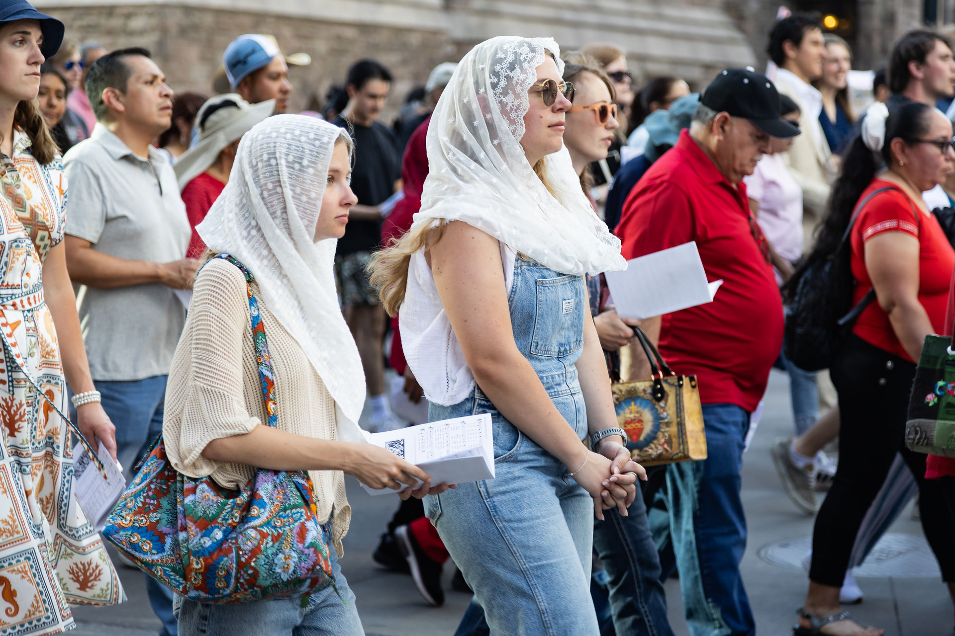 Crowd at an outdoor religious procession, with women in lace head coverings holding papers and walking solemnly.