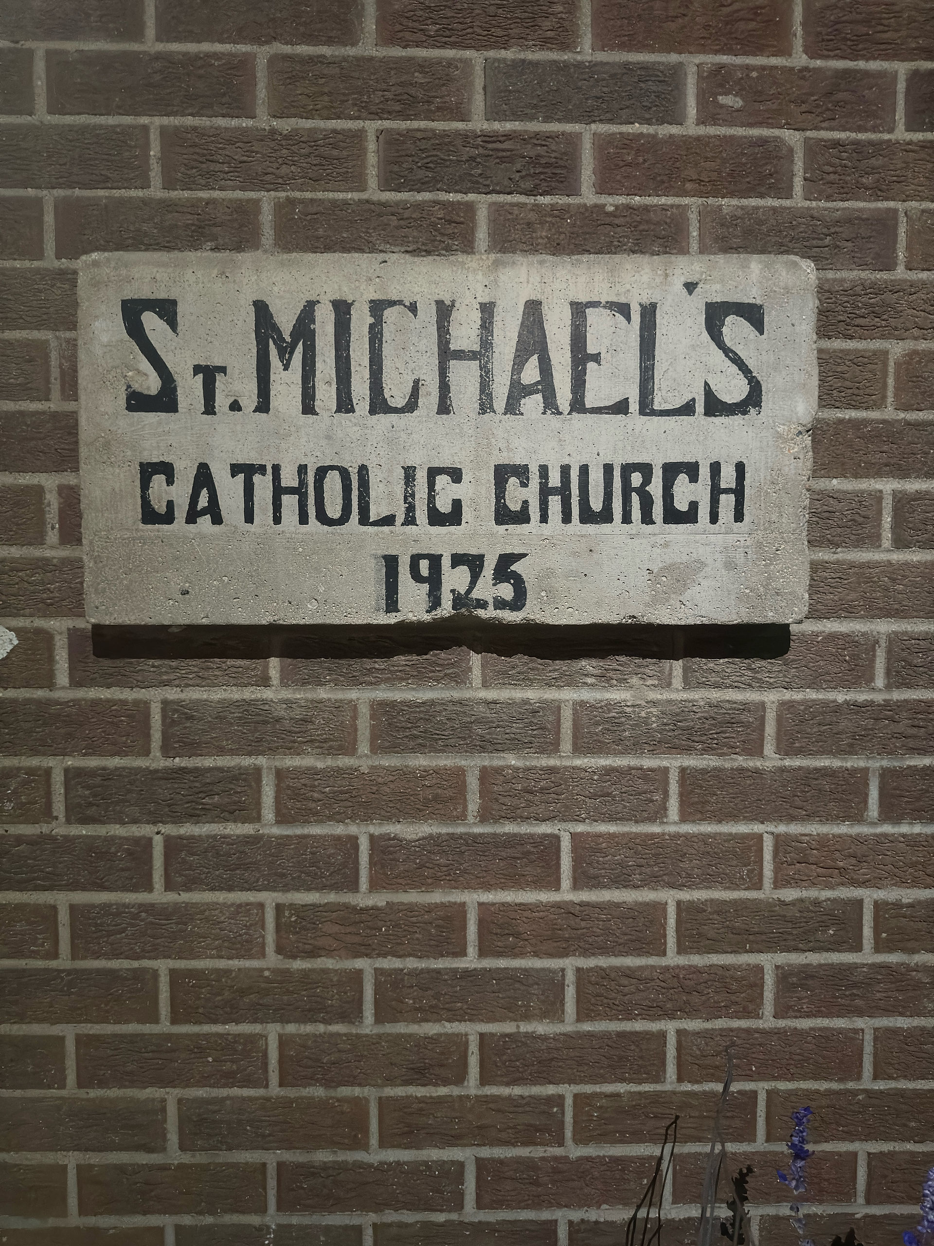 Sign reads "St. Michael's Catholic Church 1935" on a stone plaque mounted on a brown brick wall, surrounded by small purple flowers.