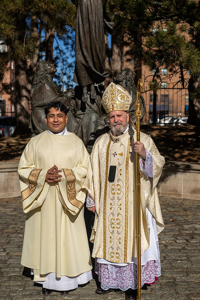 Two clergy in ornate robes pose outdoors near a statue. One holds a golden staff. Sunlit trees and buildings form the background.