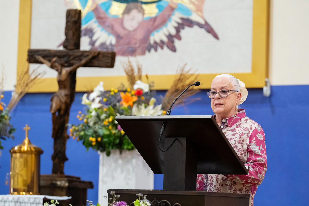 Elderly woman speaks at a lectern in a church, beside a crucifix and flowers, with a colorful angel mural in the background.