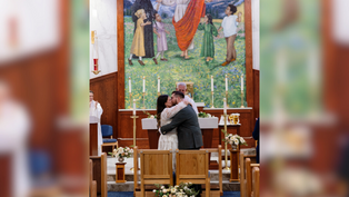 A couple kisses in a church, in front of seats and floral decor. A colorful mural is visible in the background depicting figures and nature.