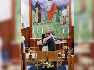 A couple kisses in a church, in front of seats and floral decor. A colorful mural is visible in the background depicting figures and nature.
