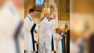 A bishop in a gold mitre performs a blessing in a church. A priest assists, while others observe. Light filters through stained glass.