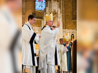 A bishop in a gold mitre performs a blessing in a church. A priest assists, while others observe. Light filters through stained glass.