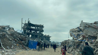 People walk through rubble-strewn street with damaged buildings. Overcast sky adds a somber mood. A child in red and a blue tarp are visible.