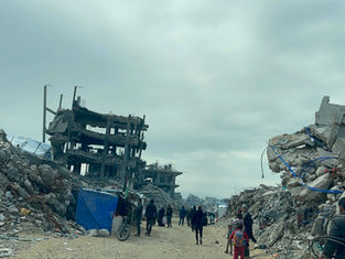 People walk through rubble-strewn street with damaged buildings. Overcast sky adds a somber mood. A child in red and a blue tarp are visible.