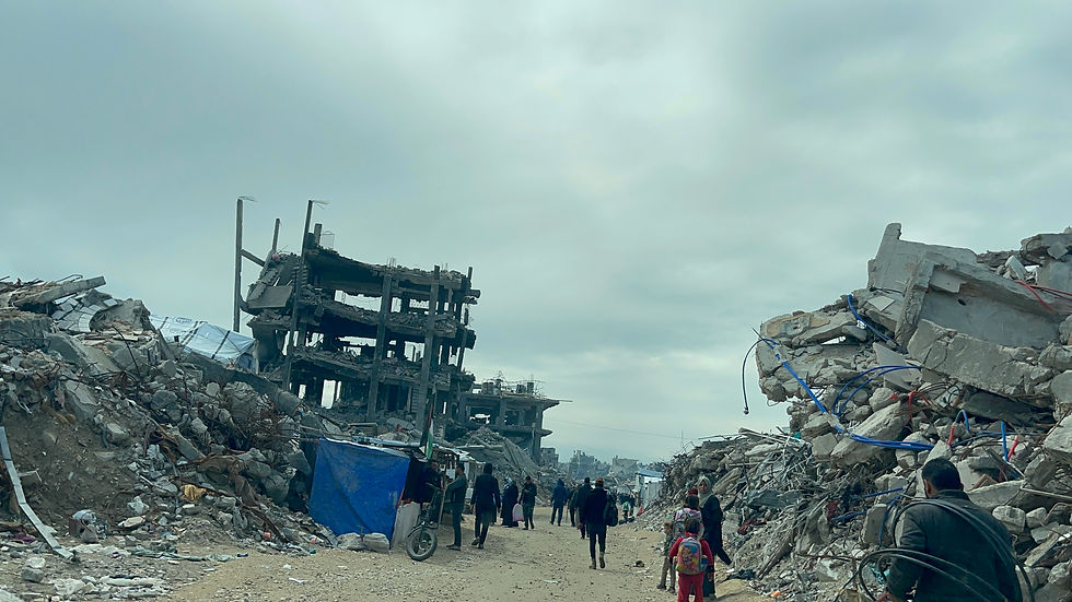 People walk through rubble-strewn street with damaged buildings. Overcast sky adds a somber mood. A child in red and a blue tarp are visible.