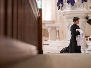 A man in black robes kneels in a church, holding white lace cloth. The interior is ornate with marble and wood, creating a solemn atmosphere.