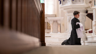 A man in black robes kneels in a church, holding white lace cloth. The interior is ornate with marble and wood, creating a solemn atmosphere.