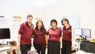 Four people in matching maroon shirts smile in an office. Desks have monitors, computers, and decor. Bright, cheerful atmosphere.