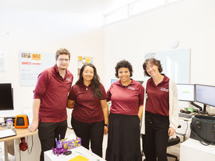 Four people in matching maroon shirts smile in an office. Desks have monitors, computers, and decor. Bright, cheerful atmosphere.