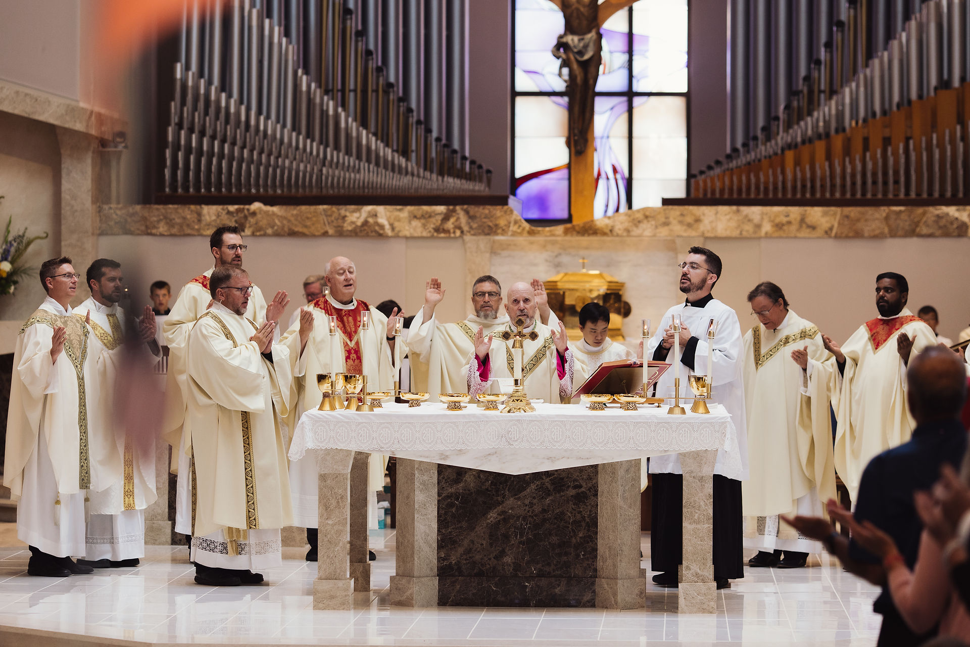 Priests in white robes conduct a religious ceremony at an altar with gold chalices. A crucifix and stained glass are in the background.
