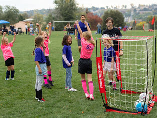 Kids cheer at a youth soccer practice on a grassy field, with a coach, mini goal net, and POWERSNET text visible on the goal.