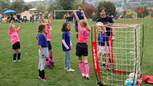 Kids cheer at a youth soccer practice on a grassy field, with a coach, mini goal net, and POWERSNET text visible on the goal.
