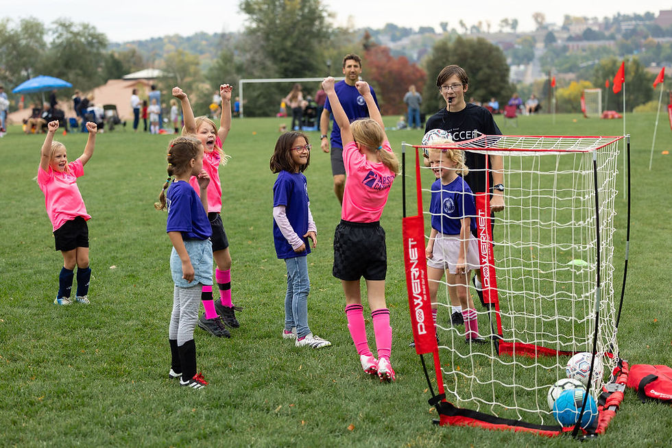 Kids cheer at a youth soccer practice on a grassy field, with a coach, mini goal net, and POWERSNET text visible on the goal.