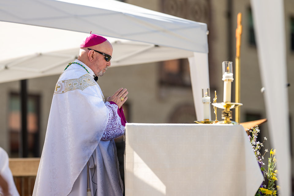 Catholic bishop in white and purple vestments prays at an outdoor altar with candles under a canopy, in a solemn ceremony