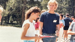 Two people talking outdoors, surrounded by others. One holds a folder. Background shows trees and bright sunlight. Casual, social setting.