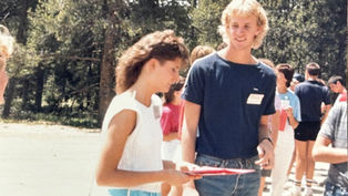 Two people talking outdoors, surrounded by others. One holds a folder. Background shows trees and bright sunlight. Casual, social setting.