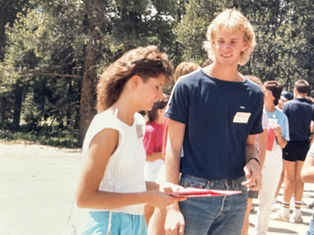 Two people talking outdoors, surrounded by others. One holds a folder. Background shows trees and bright sunlight. Casual, social setting.
