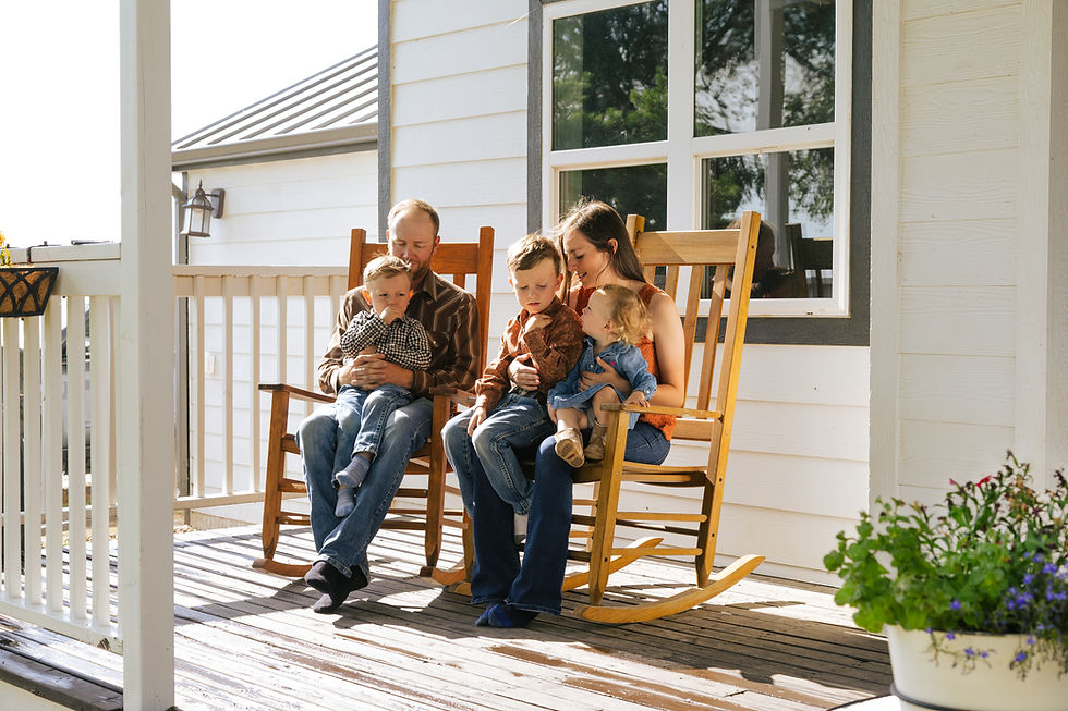 Family with two kids sitting on porch rocking chairs, white house background, sunny day. Relaxed and content mood, greenery visible.
