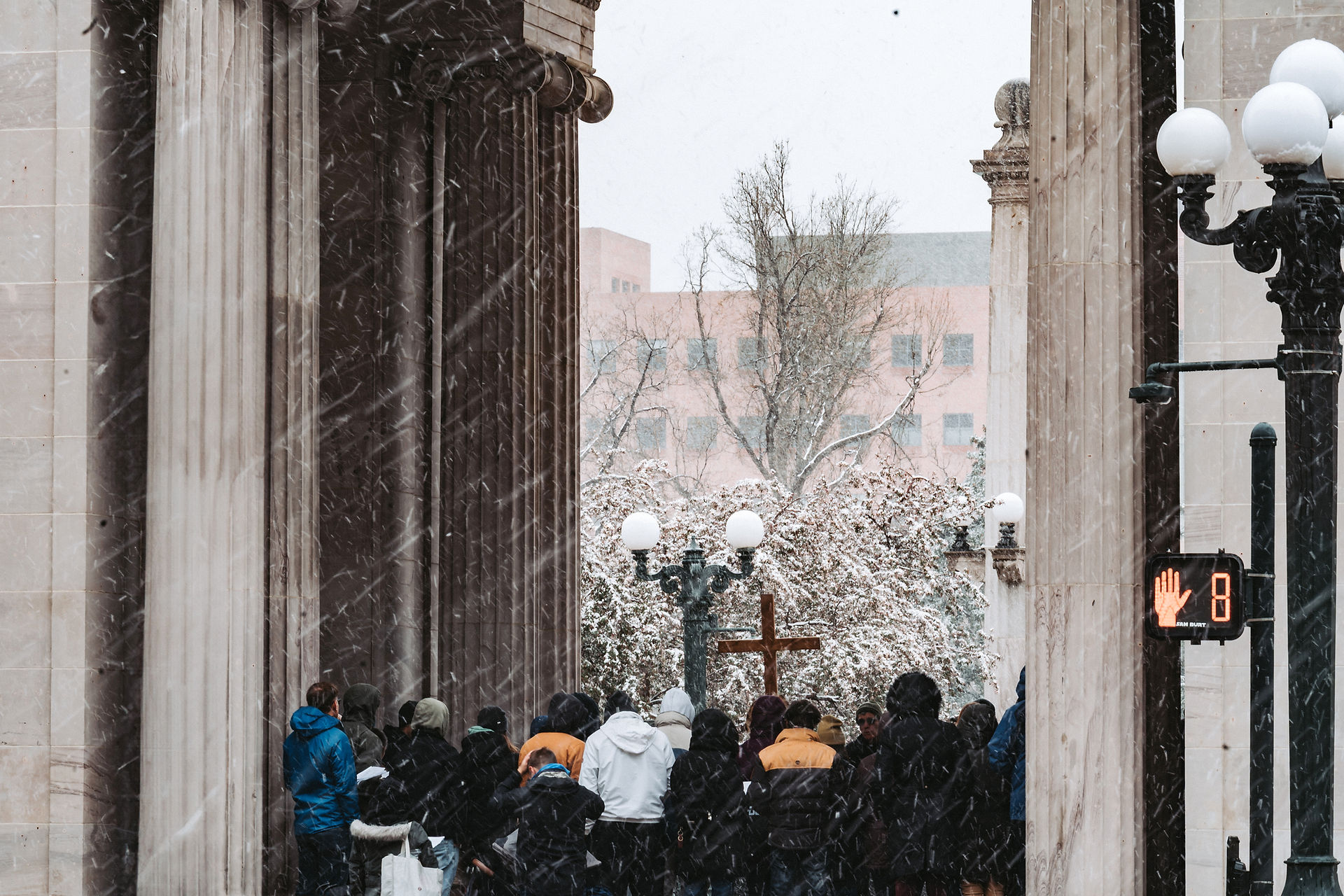 A crowd in winter coats gathers on a snowy day between tall columns. A cross is visible. Red pedestrian light shows a hand with number 8.