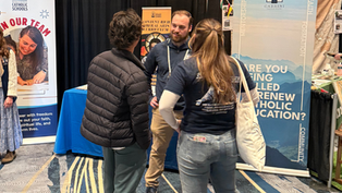 Three people converse at a booth with Catholic-themed banners. One person wears a dark jacket; others wear logo t-shirts. Colorful carpet and decor.