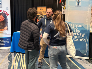 Three people converse at a booth with Catholic-themed banners. One person wears a dark jacket; others wear logo t-shirts. Colorful carpet and decor.