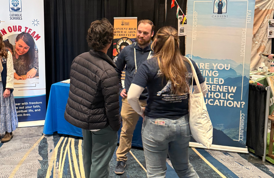 Three people converse at a booth with Catholic-themed banners. One person wears a dark jacket; others wear logo t-shirts. Colorful carpet and decor.