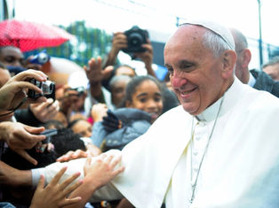 A religious figure in white attire smiles, greeting a crowd under umbrellas. Cameras capture the moment, creating a joyful atmosphere.