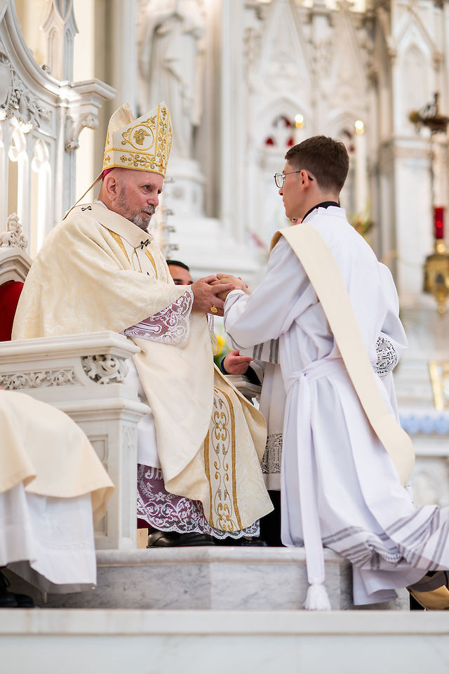 A bishop in ornate robes holds a young cleric's hands in a grand cathedral, conveying a solemn and reverent atmosphere.