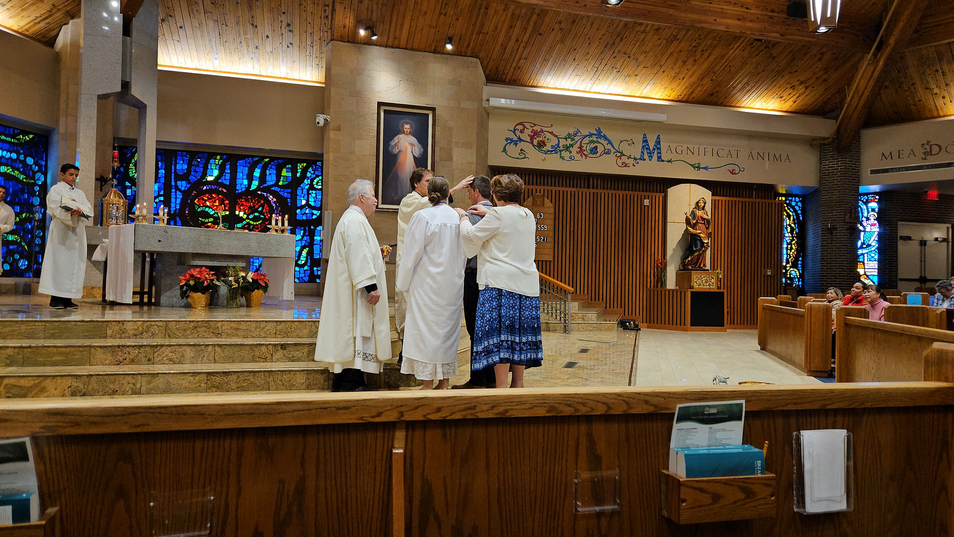 People in religious attire stand in a church with stained glass, performing a blessing. "Magnificat Anima" text is visible. Warm, solemn mood.