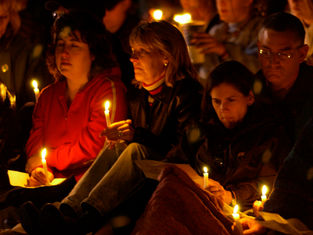 People holding candles sit closely together at a vigil, creating a somber, reflective atmosphere. Warm candlelight illuminates their faces.