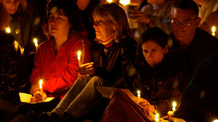 People holding candles sit closely together at a vigil, creating a somber, reflective atmosphere. Warm candlelight illuminates their faces.