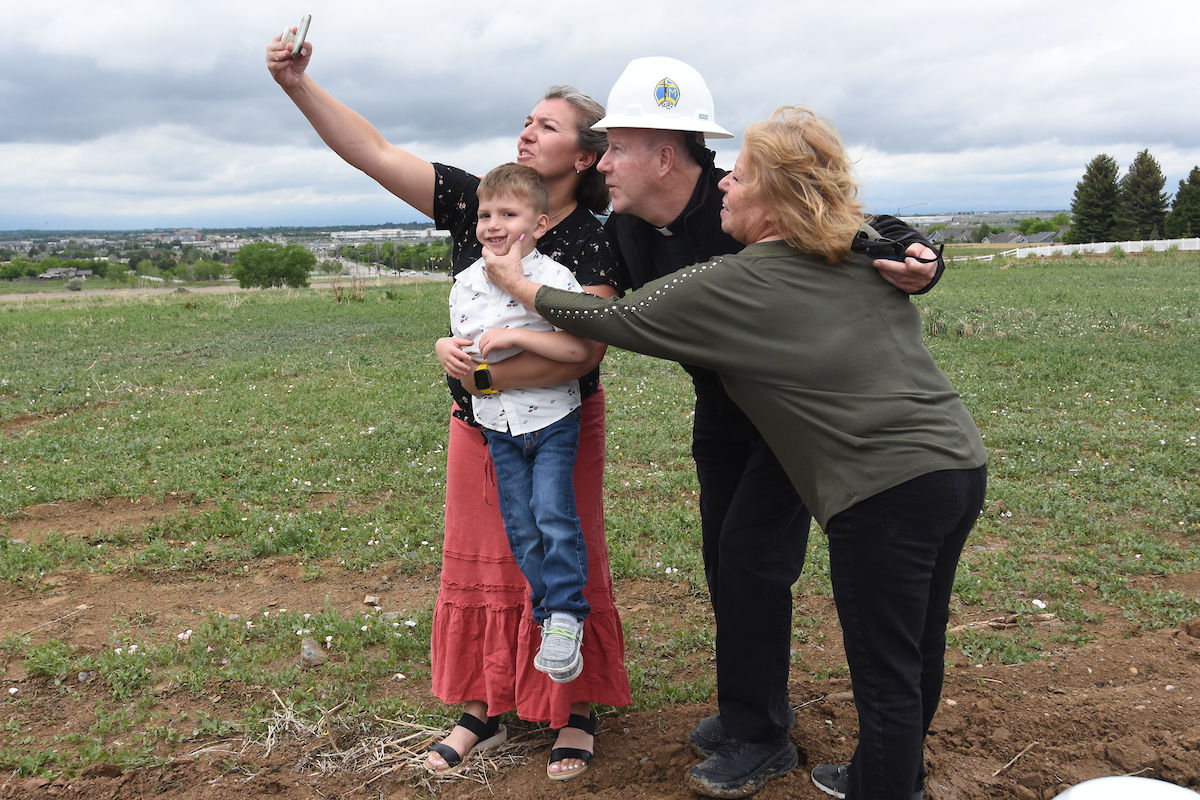A family of four takes a selfie in a grassy field. One wears a hard hat. Overcast sky. They appear happy and relaxed.