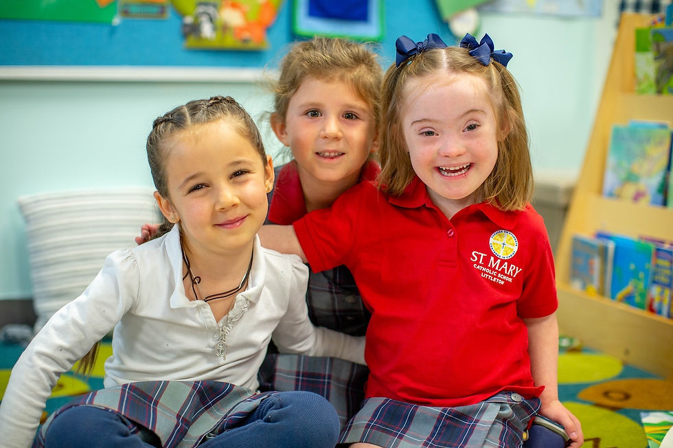 Three smiling children in a colorful classroom. One wears a red St. Mary Catholic School shirt. Books and a bulletin board are visible.