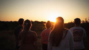 Five people stand watching a sunset in a grassy field. The sky is orange, creating a serene mood. One shirt reads "LIFE is better".