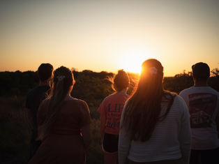 Five people stand watching a sunset in a grassy field. The sky is orange, creating a serene mood. One shirt reads "LIFE is better".