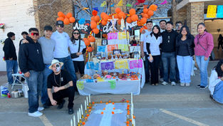 A group poses by a colorful Day of the Dead altar with orange balloons, photos, and candles. Outdoors, sunny, festive mood.