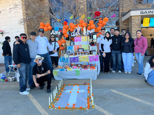 A group poses by a colorful Day of the Dead altar with orange balloons, photos, and candles. Outdoors, sunny, festive mood.