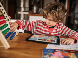 Child in a red-striped shirt using an abacus and tablet at a wooden table, focused and engaged. Books and papers are nearby.