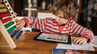 Child in a red-striped shirt using an abacus and tablet at a wooden table, focused and engaged. Books and papers are nearby.