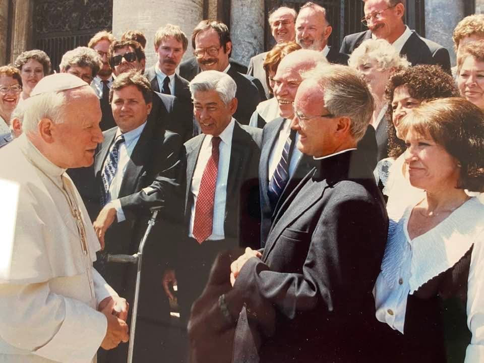 Pope John Paul II talks with smiling clergy and officials outdoors on stone steps in bright sunlight.