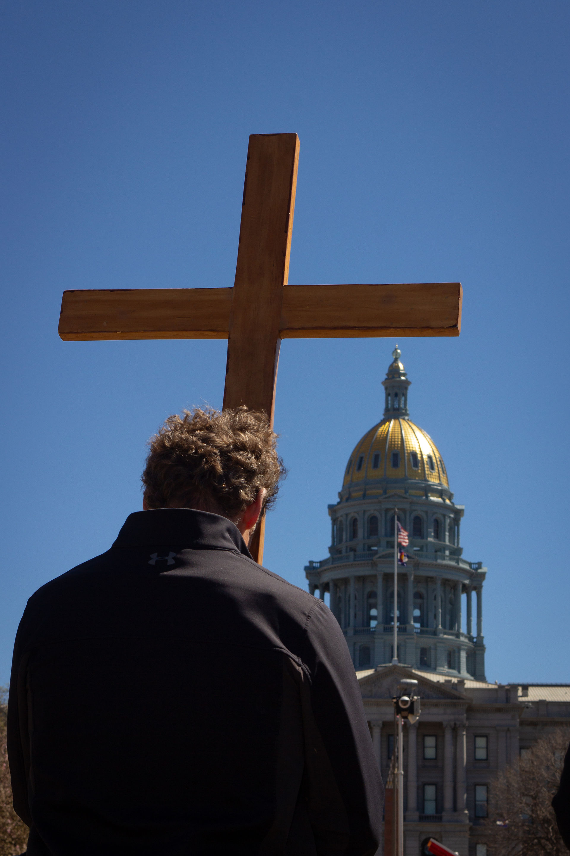A man carries a large wooden cross while facing the Colorado State Capitol building under a clear blue sky.