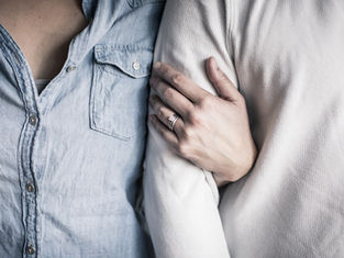 Person in denim shirt stands next to another in white, with an arm linked and hand displaying rings, creating an intimate, close atmosphere.