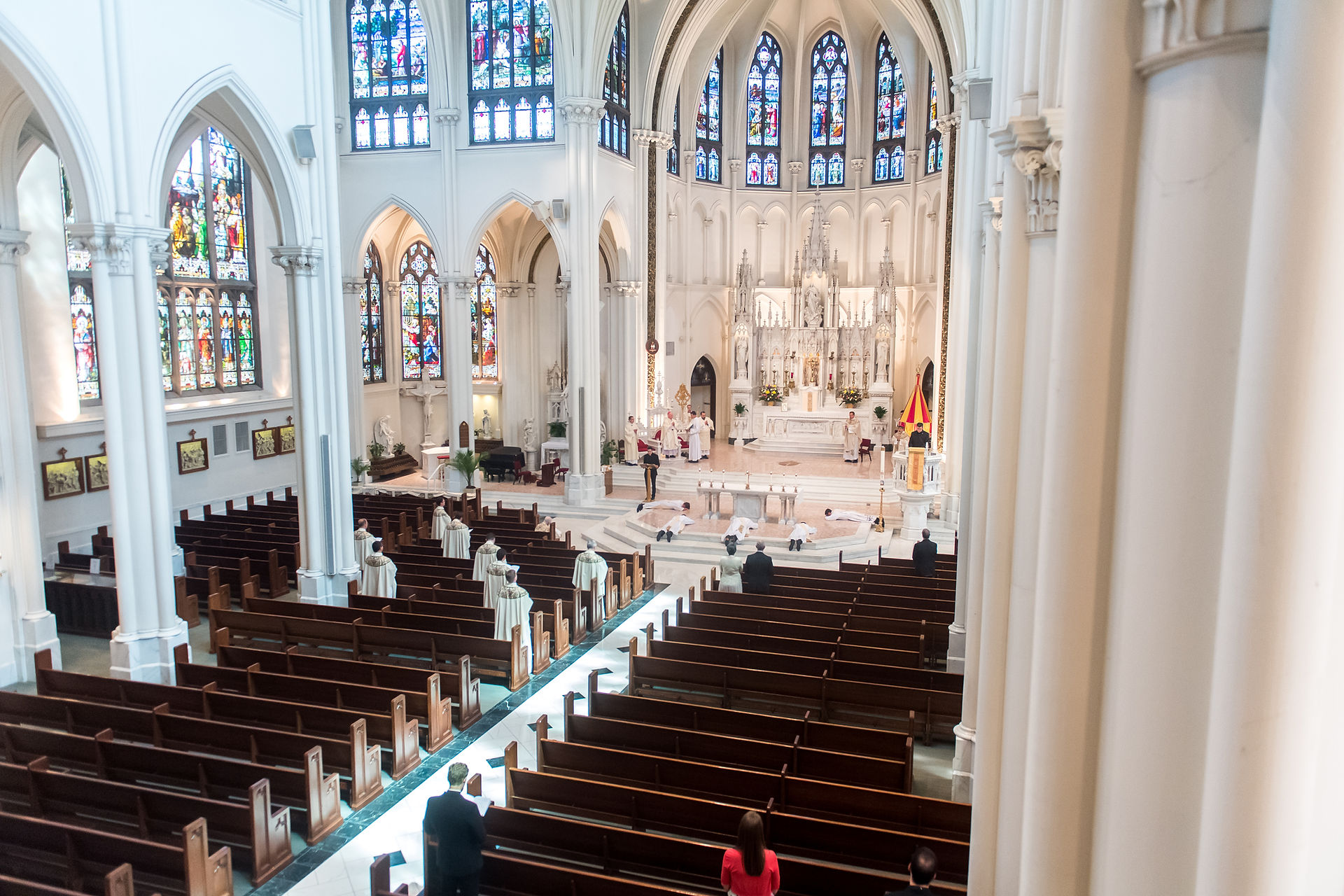 Spacious church interior with stained glass windows. People in robes stand near the altar, some kneel. Empty wooden pews create a serene mood.