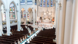 Wide view of a cathedral interior with stained glass windows. Clergy in robes perform a ceremony at the altar, while people observe from pews.